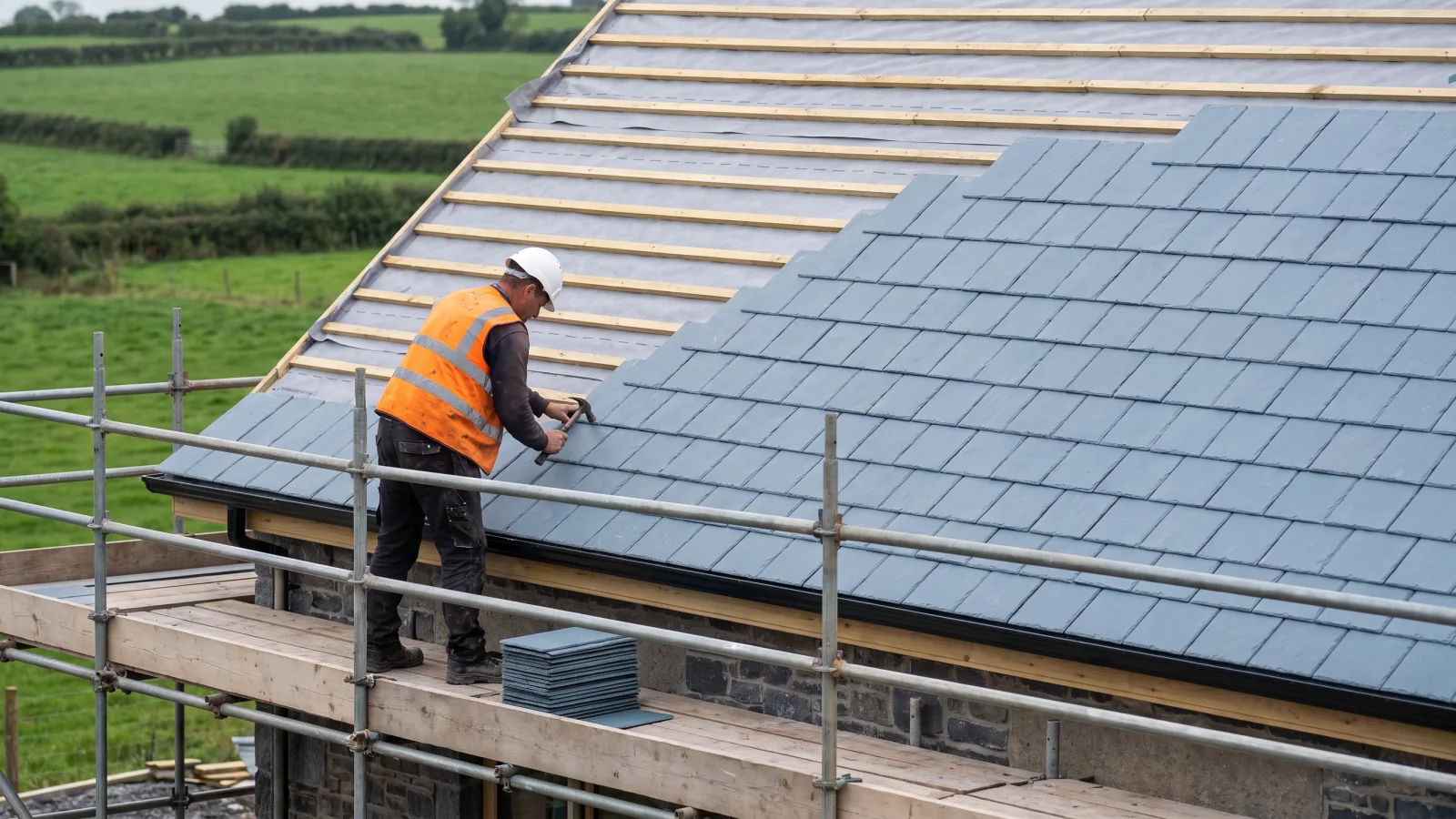A roofer in a hard hat, hi-vis vest and safety boots fixing slates from a scaffold with guardrails, a stack of slates on the boards